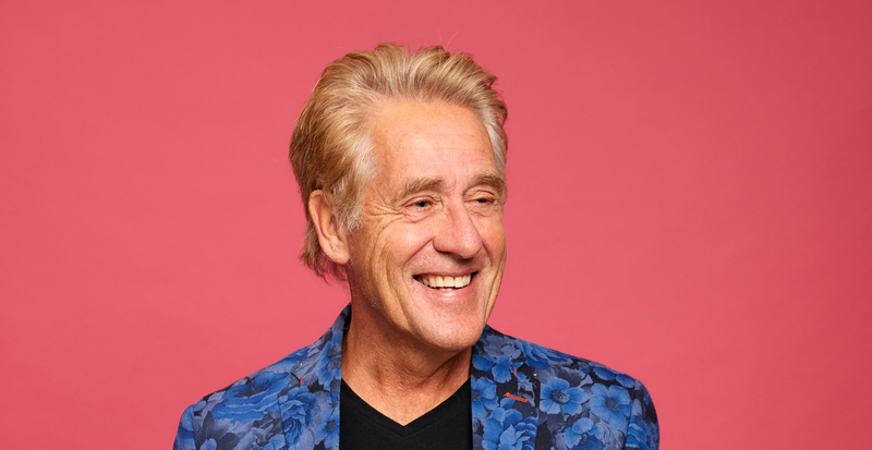 Man met een glimlach en een kleurrijke blazer poseert voor een roze achtergrond.