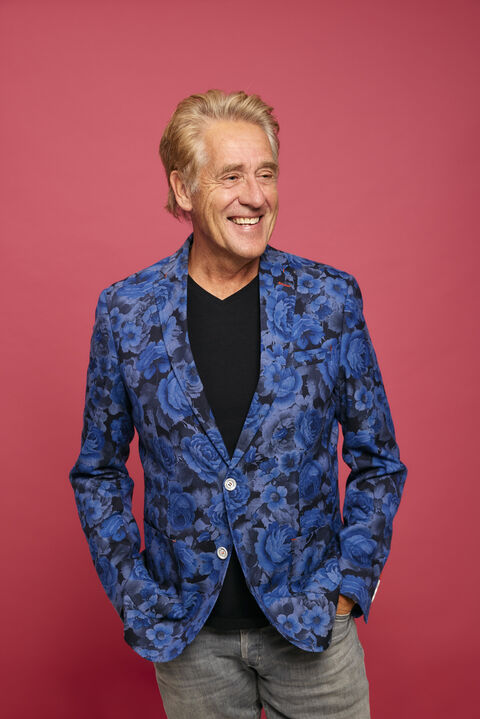Man met een grijs-blonde haarkleur in een blauwe blazer met bloemenpatroon, smiling tegen een roze achtergrond.