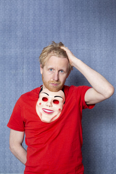Man met een rood T-shirt en een maskersculptuur op zijn borst staat met de hand in zijn haar voor een blauwe achtergrond.
