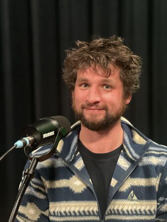 Een man met krullend haar en een baard, gekleed in een gestreepte trui, staat bij een microfoon en kijkt naar de camera.