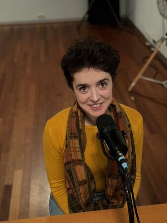 Vrouw met een donkere krullende haar, in een gele trui en sjaal, kijkt lachend recht in de camera terwijl ze voor een microfoon zit.
