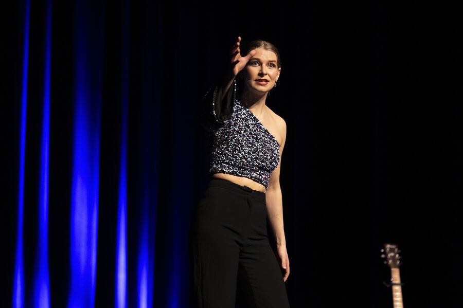 Een jonge vrouw in een blauwe top en zwarte broek staat op het podium en spreekt naar het publiek met een gitaar naast haar.