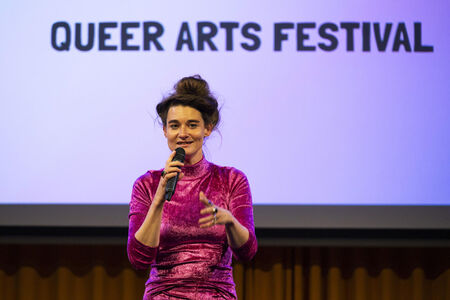 Vrouw in een glinsterende roze outfit spreekt in een microfoon voor een scherm met de tekst "QUEER ARTS FESTIVAL".