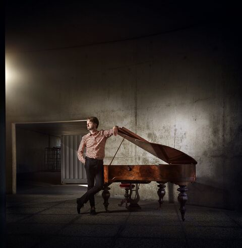 Man leunt tegen een openstaande piano in een donkere, industriële ruimte met een betonnen muur.