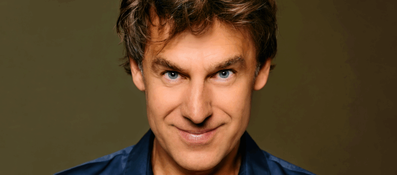 Portret van een man met een vrolijke uitstraling en stekelig bruin haar, tegen een eenvoudige achtergrond.