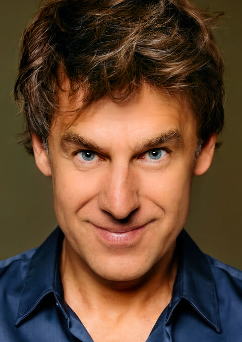 Portret van een man met bruin haar en blauwe ogen, glimlachend in een zwarte blouse tegen een neutrale achtergrond.