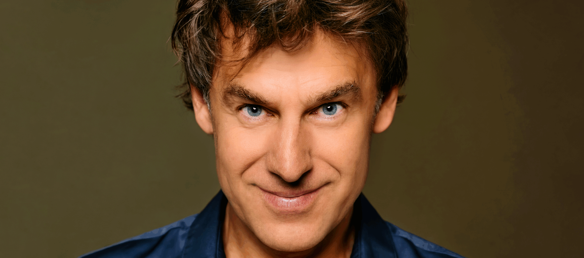 Portret van een man met een vrolijke uitstraling en stekelig bruin haar, tegen een eenvoudige achtergrond.