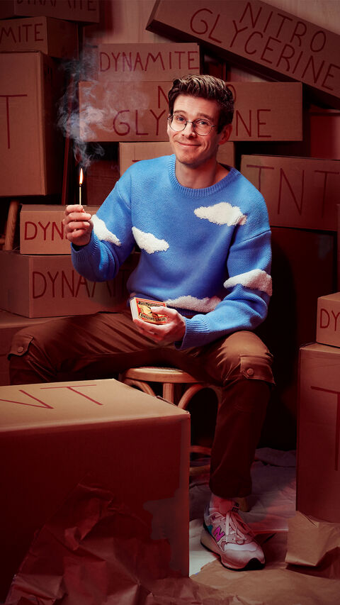 Man in een blauwe trui met wolkenprint zit tussen dozen en houdt een aansteker vast. Achtergrond is vol met dynamietdozen.