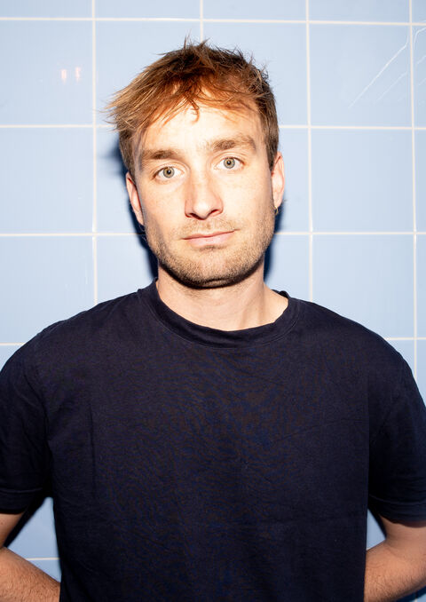 Portret van een man met kort, lichtbruin haar, staand voor een blauwe tegelwand, met een neutrale gezichtsuitdrukking.