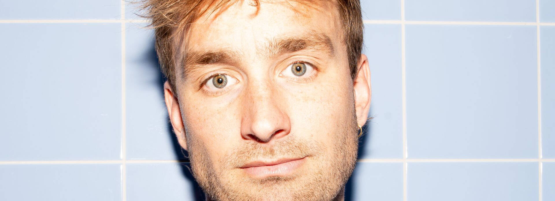 Een portret van een man met blond haar, een neutrale gezichtsuitdrukking, tegen een lichtblauwe achtergrond.