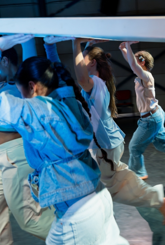Groep dansers beweegt dynamisch onder een horizontale structuur in een repetitieve oefening.