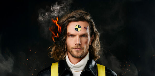 Man met lang haar en een crash-test symbool op zijn voorhoofd, gefotografeerd met een brandende achtergrond.