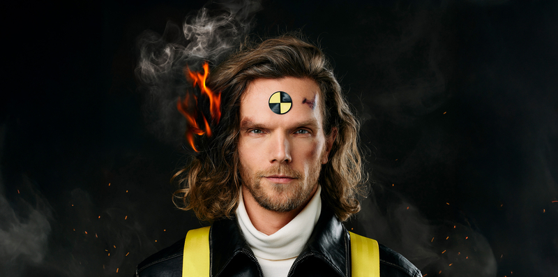 Man met lang haar en een crash-test symbool op zijn voorhoofd, gefotografeerd met een brandende achtergrond.