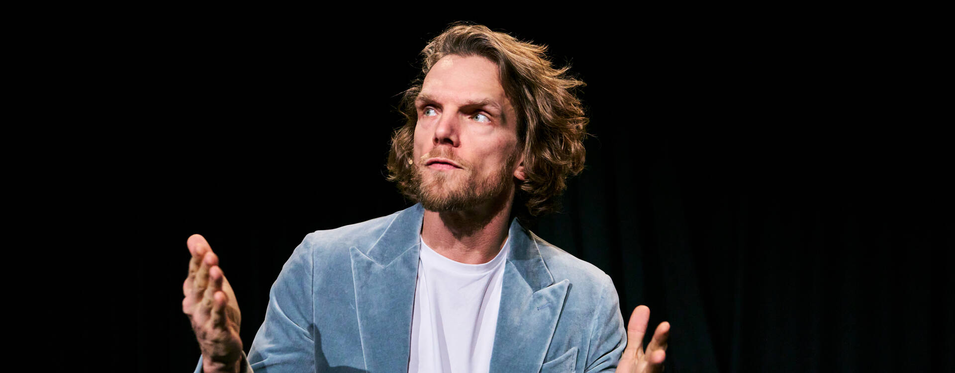 Man in lichtgekleurde blazer met een serieuze uitdrukking, beide handen omhoog, op een donkere achtergrond.