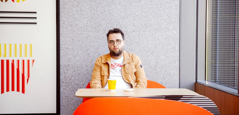 Man met een beige jas zit aan een oranje tafel met een gele beker in een moderne, lichte ruimte.