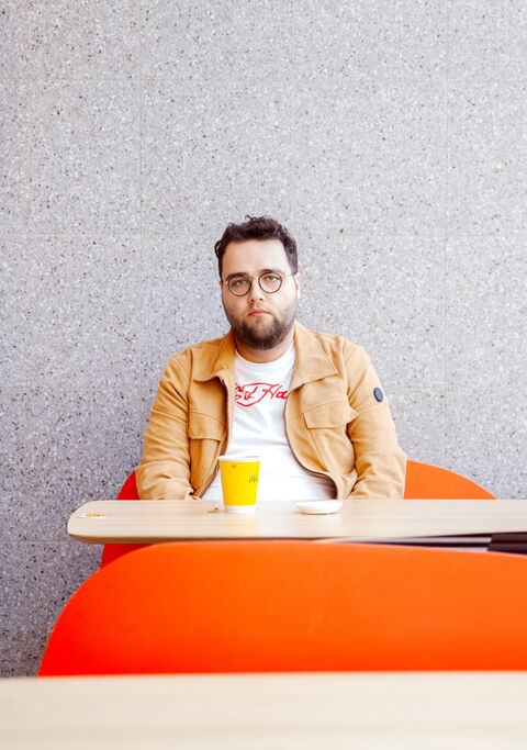 Man met een bril en een beige jas zit aan een tafel met een oranje blad, houdt een geel drankje vast voor een grijze muur.