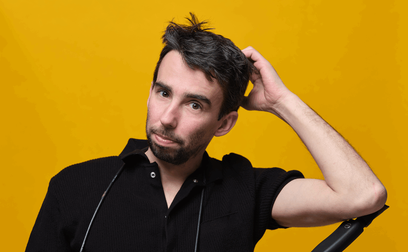 Man met kort, warrig haar en een neutrale uitdrukking, staand voor een gele achtergrond.