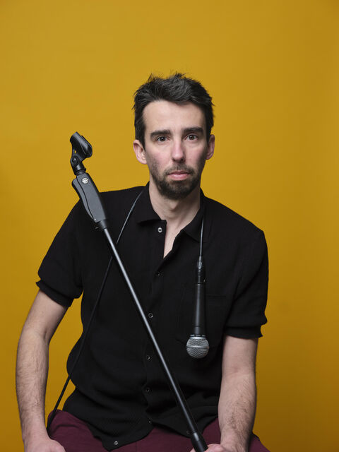 Man met een microphone en een wandelstok, zittend voor een gele achtergrond.