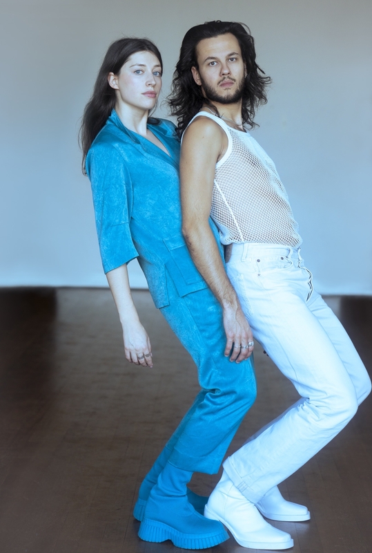 Twee mensen staan met hun ruggen tegen elkaar in een minimalistische ruimte, met modieuze outfit in blauw en wit.