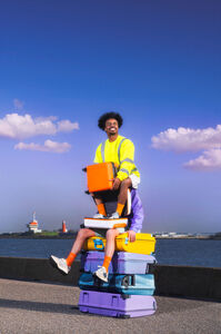 Een persoon in een veiligheidsvest zit op een stapel kleurrijke koffers aan de kust, met een heldere blauwe lucht op de achtergrond.