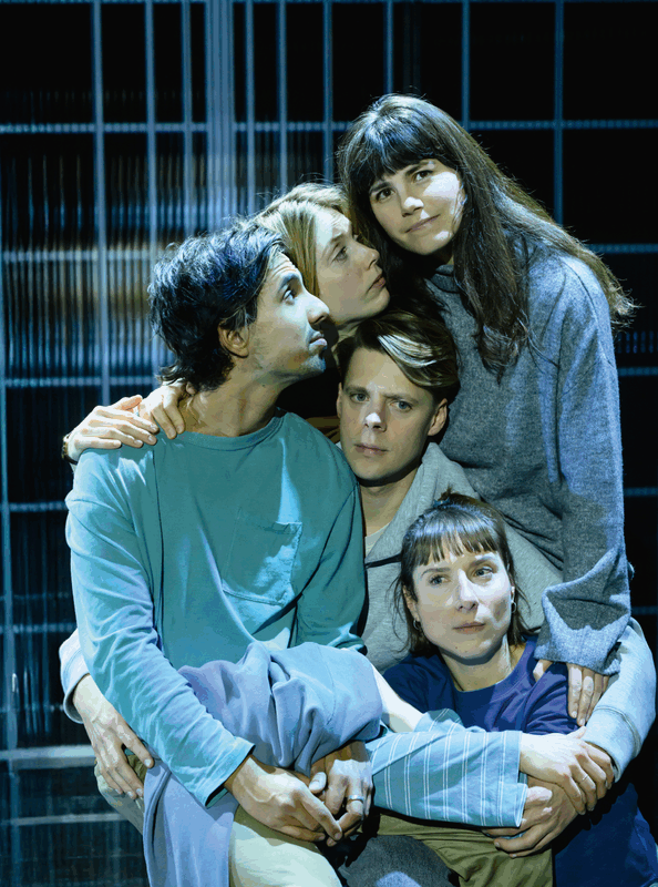 Groep van vijf mensen in een theatrale pose, zittend tegen een achtergrond van verticale lijnen, met een emotionele uitdrukking.
