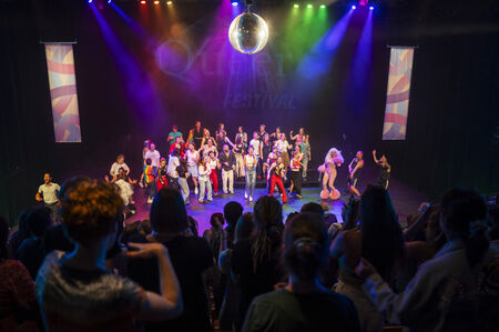 Groep mensen in kleurrijke kostuums op een podium met discobal en sfeerverlichting tijdens een evenement.