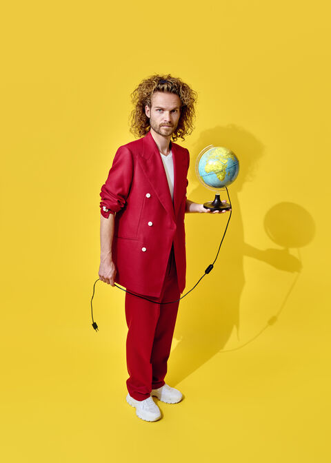 Man in een felrode outfit houdt een globe vast en staat voor een gele achtergrond.
