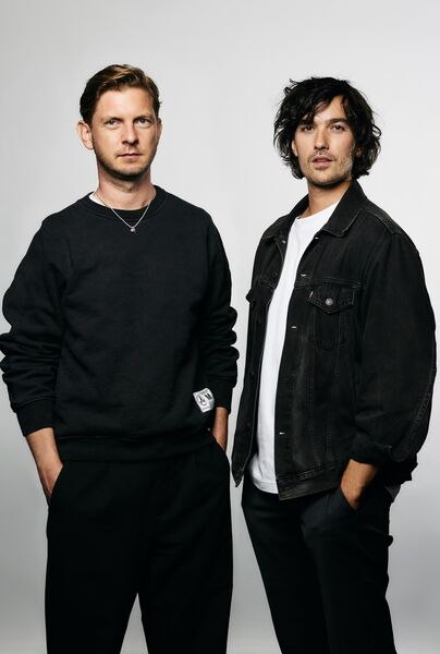 Twee jonge mannen poseren samen voor een neutrale achtergrond, met casual kleding en ontspannen houdingen.