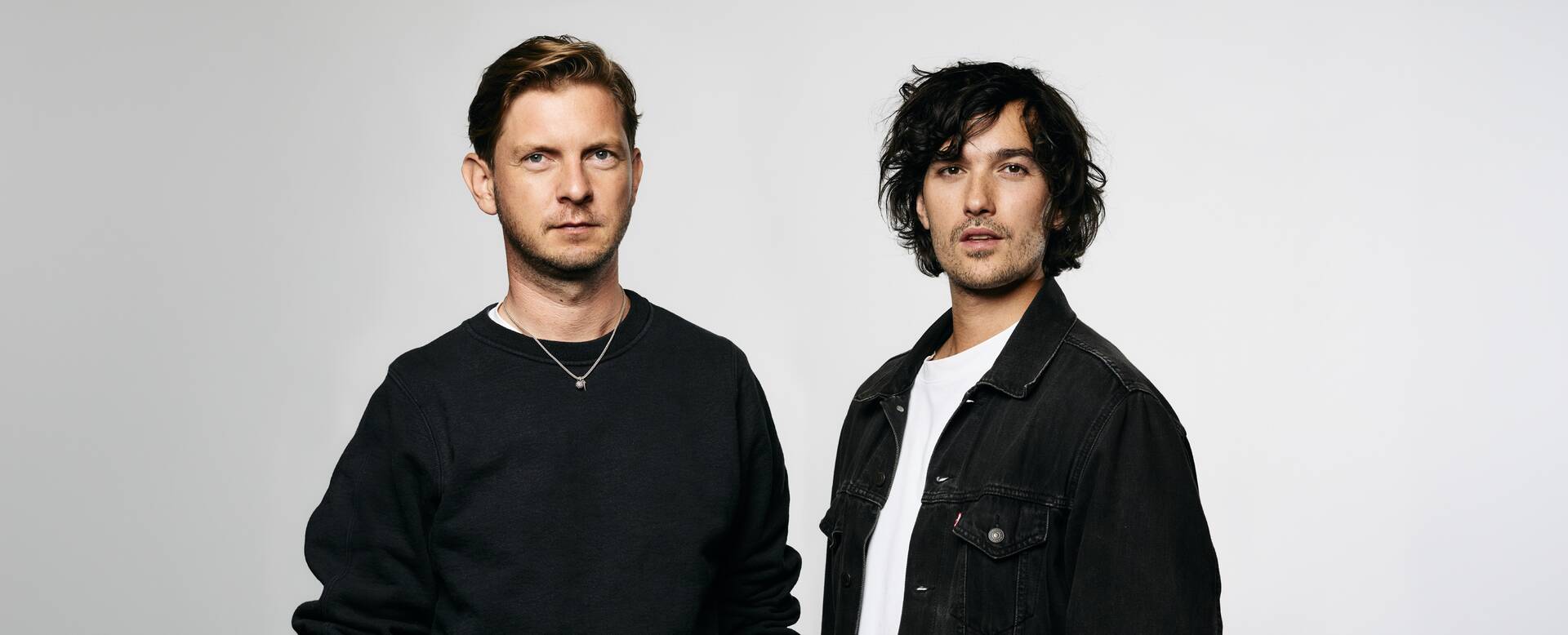 Twee mannen staan naast elkaar in een neutrale achtergrond, beiden gekleed in casual zwarte en witte kleding.