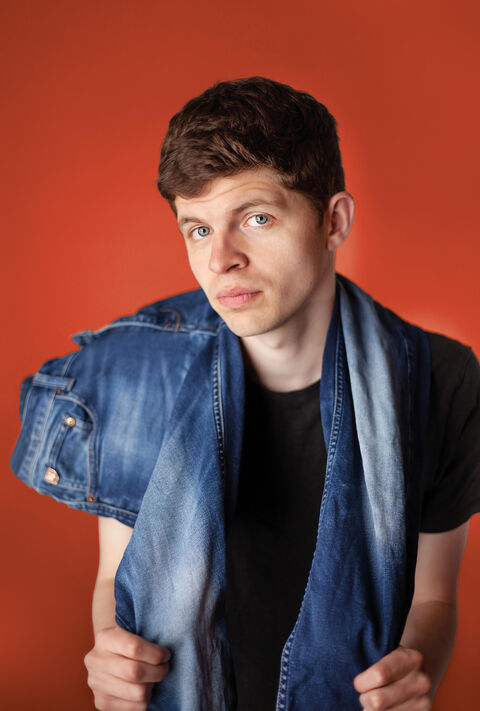 Jongeman poseert met een spijkerjasje over zijn schouder voor een oranje achtergrond.