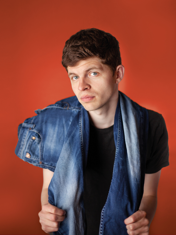 Jongeman met kort, bruin haar, draagt een zwart T-shirt en een spijkerjack op een oranje achtergrond.