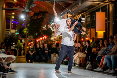 Dansvoorstelling met een performer die een andere danser in de lucht tilt, omringd door een publiek in een moderne setting.