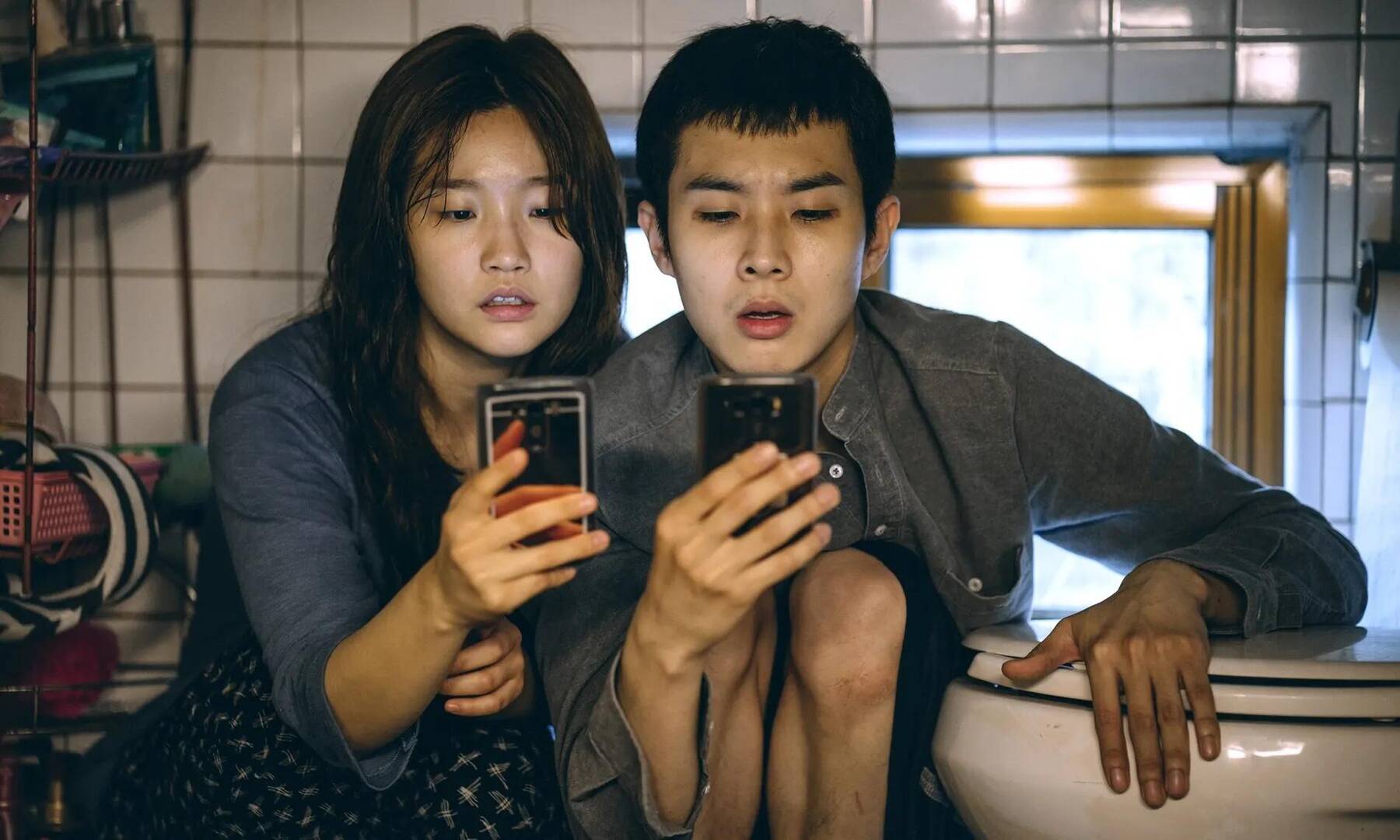 Twee jonge mensen zitten naast elkaar in een badkamer en kijken aandachtig naar hun smartphones.