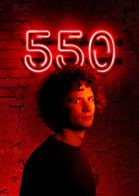 Man met krullend haar staat voor een muur, met de neonverlichting '550' boven hem.
