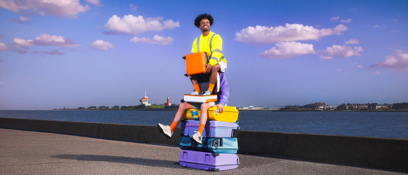 Een persoon in een felgekleurde outfit zit op een stapel koffers aan de waterkant onder een heldere lucht.