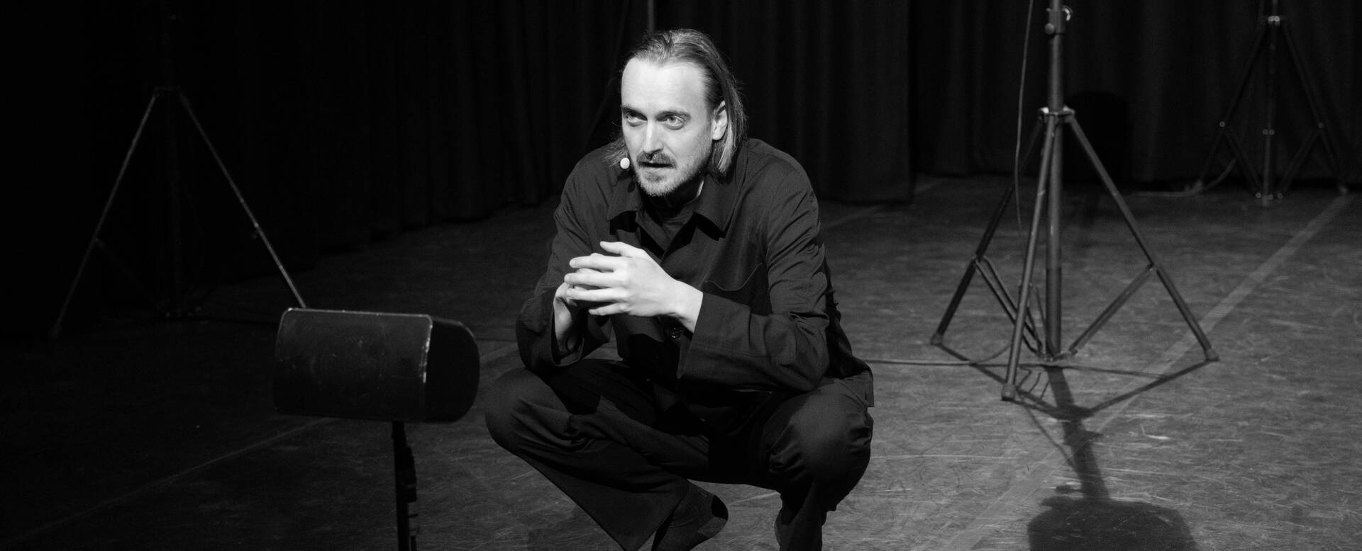 Zwart-wit afbeelding van een man die in een theateromgeving zit, met een microfoon en statief in de achtergrond.