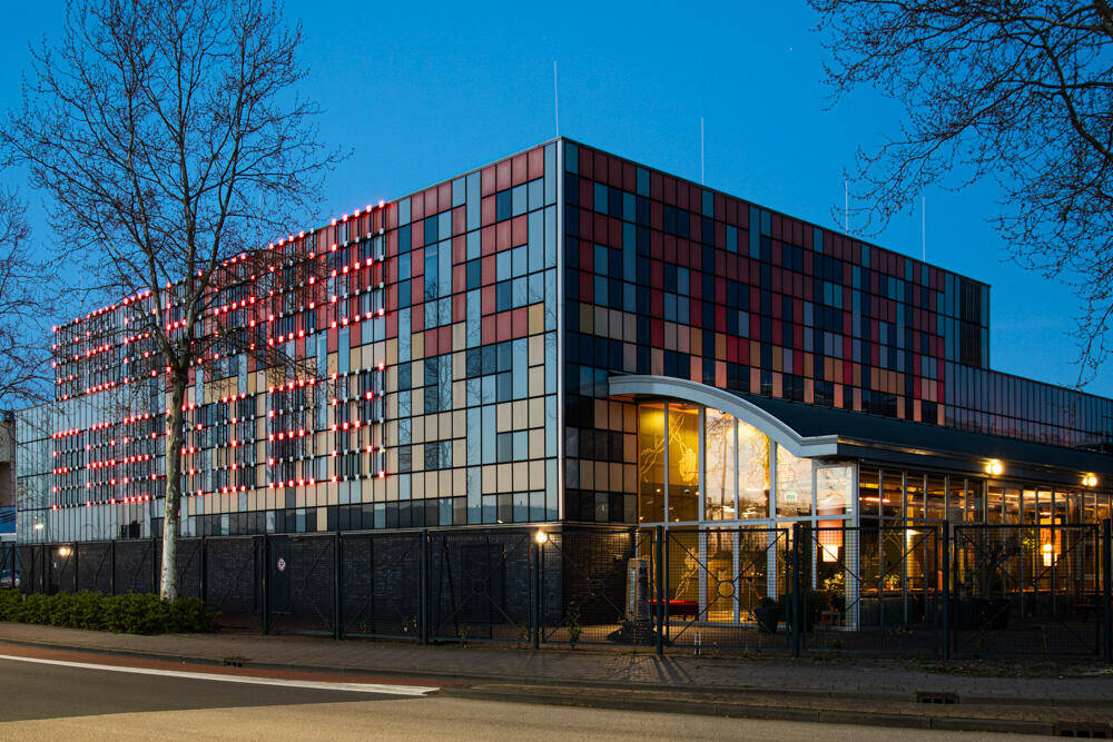 Moderne gebouw met een kleurrijke façade, verlicht in de schemering, omringd door bomen en een straat.