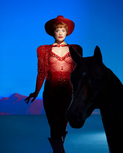 Een vrouw in een opvallende rode outfit en hoed poseert naast een zwart paard, met een abstracte blauwe achtergrond.