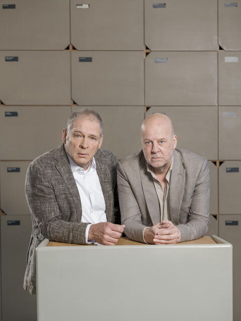 Twee mannen zitten aan een tafel voor een wand met opbergkasten, beiden gekleed in grijze jassen.
