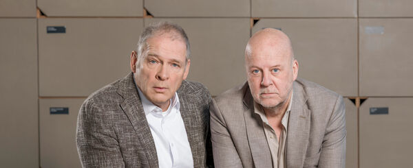Twee mannen in zakelijke kleding zitten voor een achtergrond van dozen en kijken serieus in de camera.