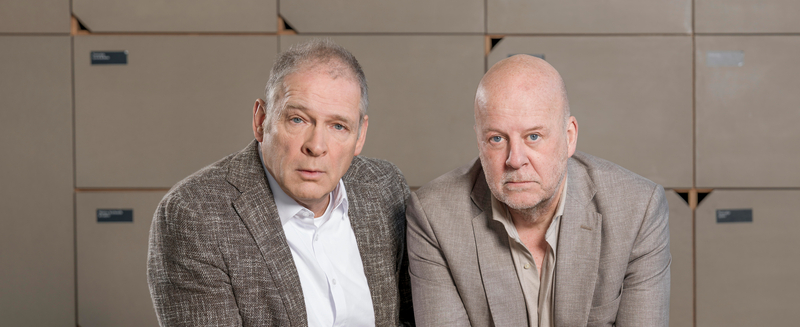 Twee mannen in zakelijke kleding zitten voor een achtergrond van dozen en kijken serieus in de camera.
