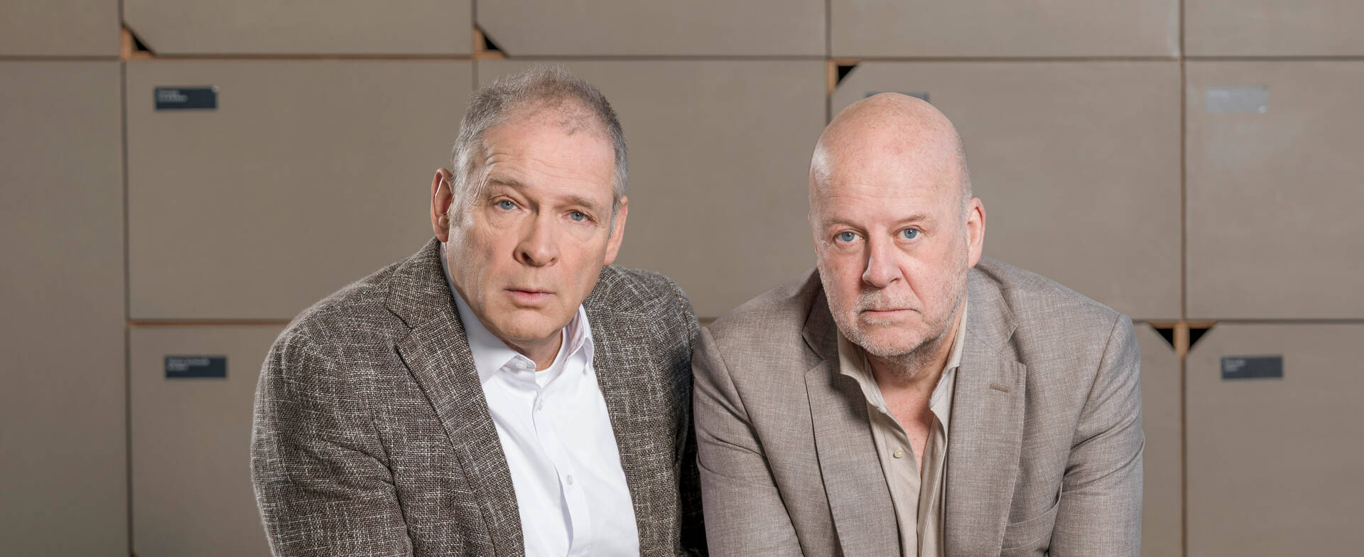 Twee mannen in zakelijke kleding zitten voor een achtergrond van dozen en kijken serieus in de camera.