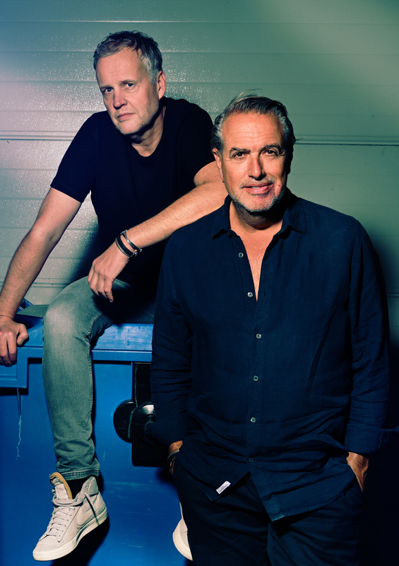 Twee mannen poseren voor een blauwe achtergrond, één zittend en de ander staand, beiden met een ontspannen uitstraling.