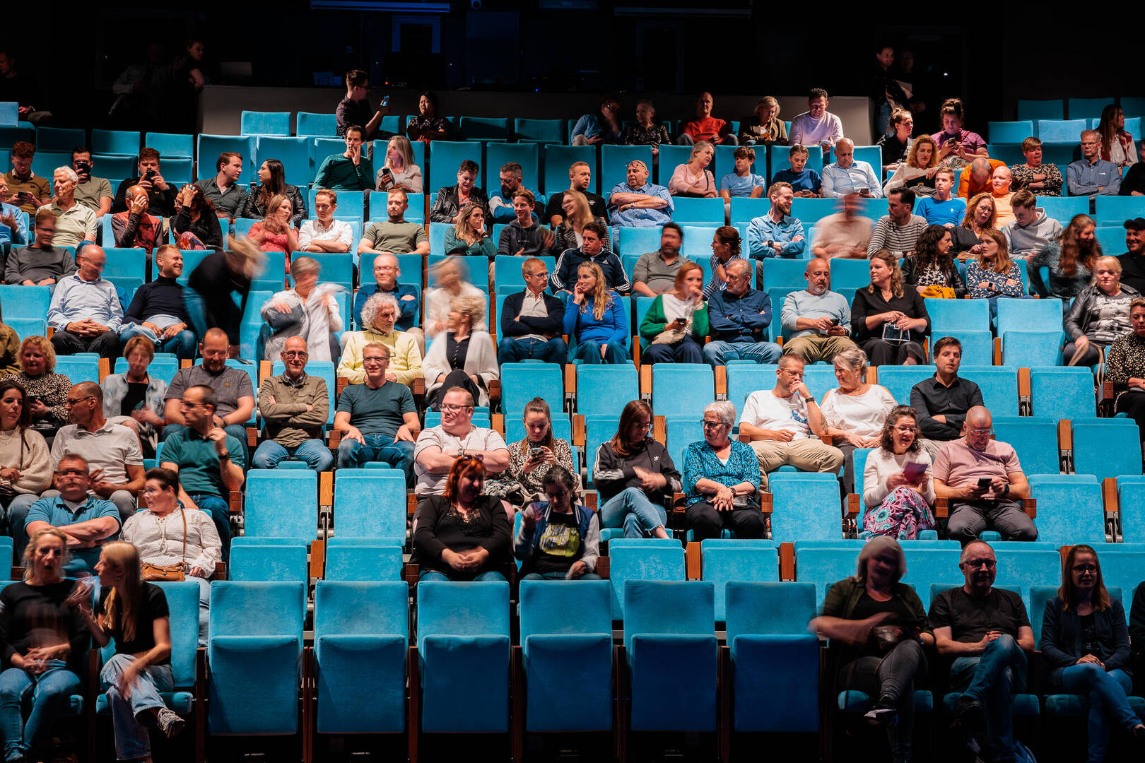 Een theaterzaal met blauwe stoelen en een publiek dat wacht op een voorstelling.