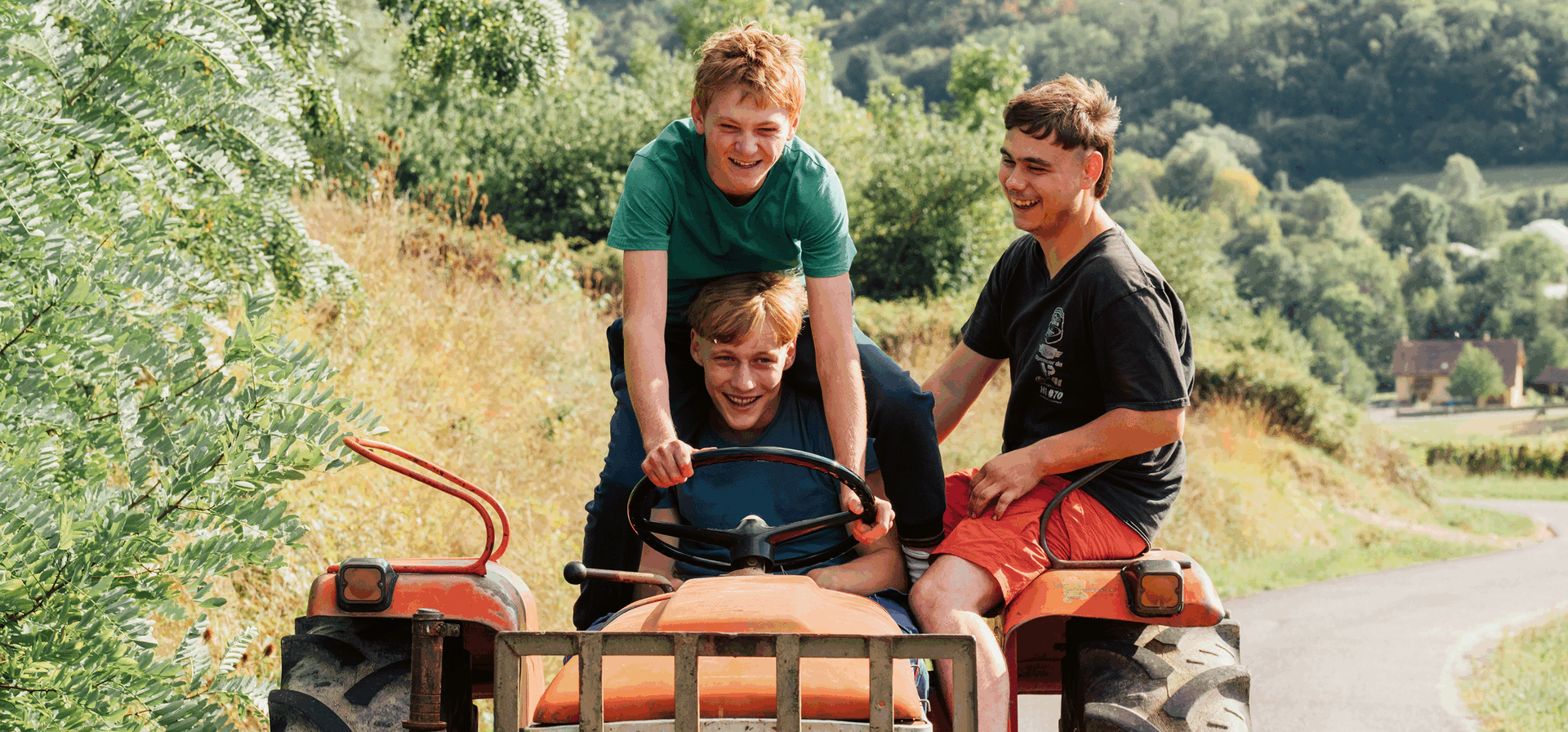 Drie tieners staan en zitten op een tractor, rijden op een landweg omgeven door natuur en heuvels.