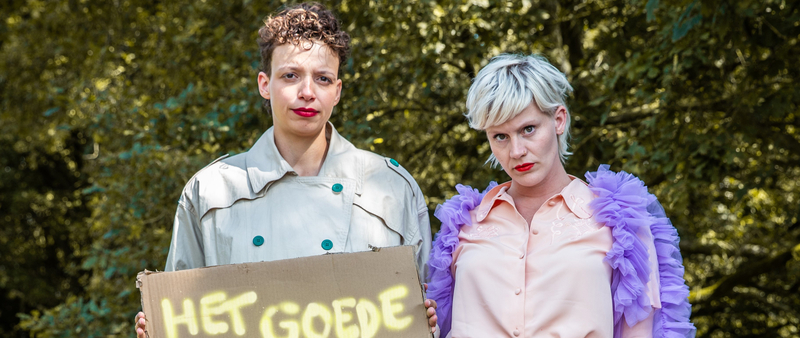 Twee mensen staan in een buitenomgeving, met een bord dat "HET GOEDE" zegt. Ze dragen opvallende kleding en maken een statement.