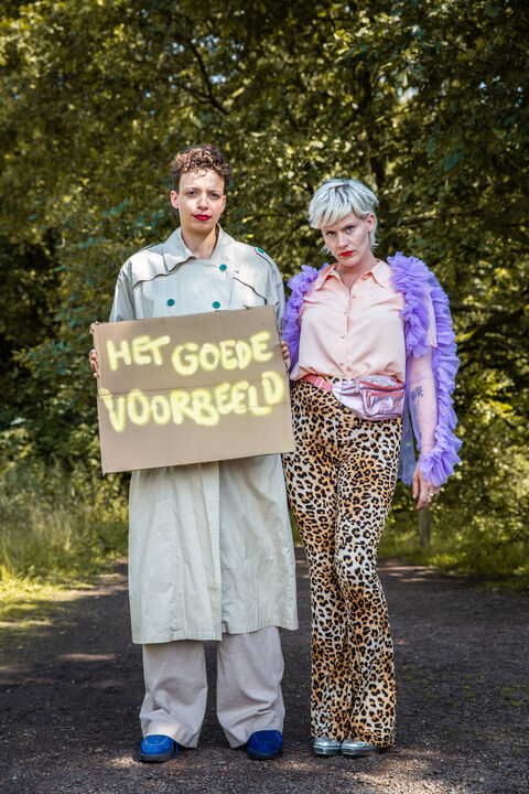 Twee performers in kleurrijke kostuums staan op een pad, met een bord waarop "Het goede voorbeeld" staat.