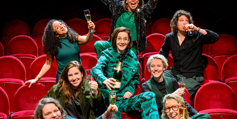 Groep vrouwen in een theaterzaal, lachend en poseerend met drankjes, in feestelijke setting.
