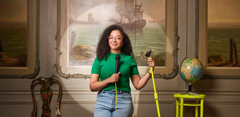 Vrouw met krullend haar in groene shirt houdt een tuinslang vast, voor een schilderij en globe in een klaslokaal.