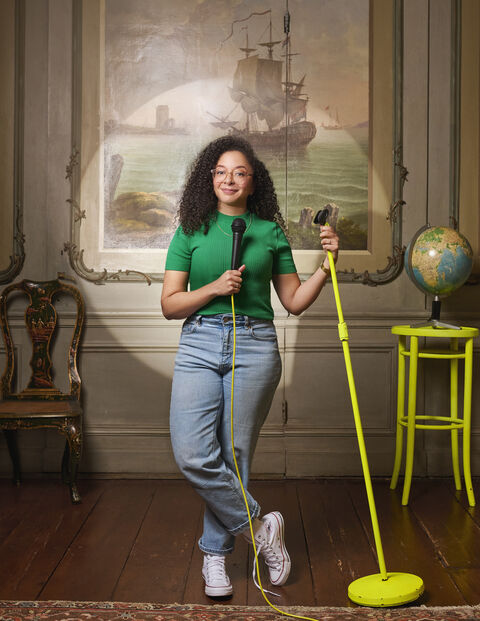 Vrouw met krullend haar in groene shirt en jeans, houdt een microfoon en een poetsapparaat vast in een elegante kamer.